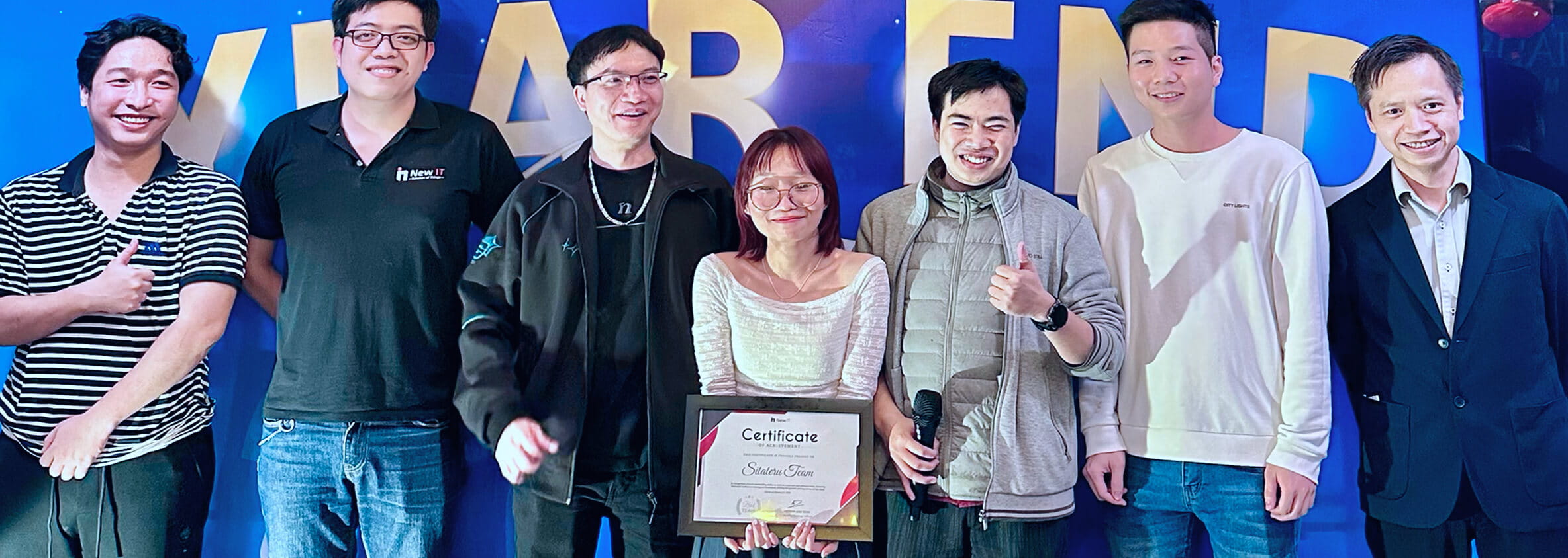 7人の男女が並んで笑顔で記念撮影している。中央の女性は「Certificate」と書かれた賞状を持っている。