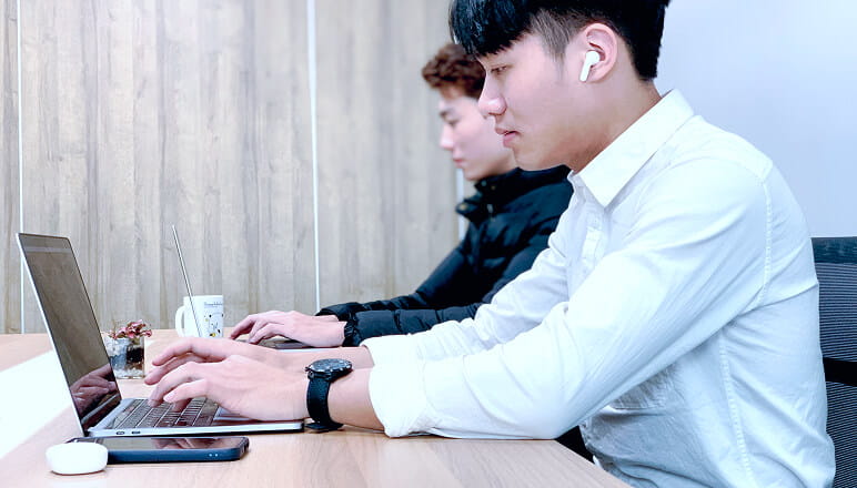 白いシャツの男性がノートパソコンで作業しながらイヤホンを装着している。